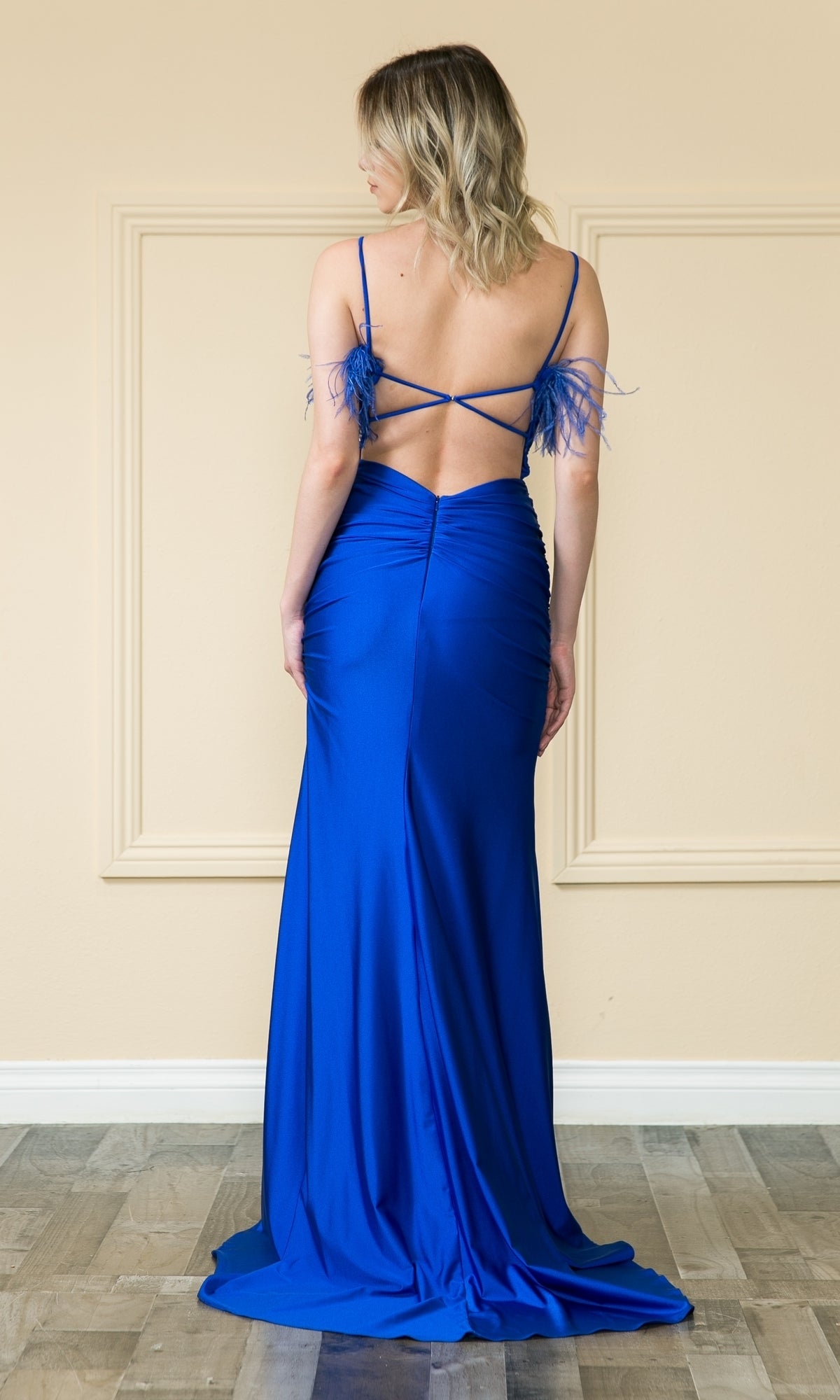 A woman stands facing away in the affordable Open-Back Long Feathered Prom Dress 9070—a royal blue, floor-length gown with thin straps, a low open back, and feathered accents on the shoulders.